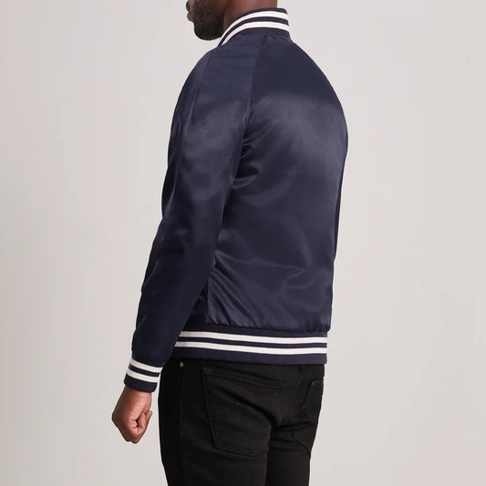 A man wearing a navy blue satin bomber jacket with white and navy striped ribbed collar, cuffs, and hem, shown from a side-back angle against a light gray studio background.
