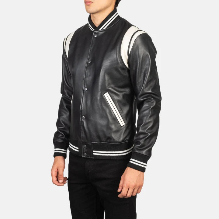 A man wearing a black leather varsity-style jacket with white contrast panels on the shoulders and sleeves, and white stripes on the collar, cuffs, and hem, paired with black pants against a light gray background.