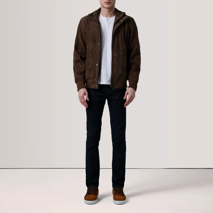 A man wearing a brown suede hooded jacket over a white t-shirt, black jeans, and brown suede shoes, standing against a plain off-white studio background.