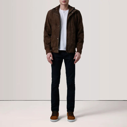 A man wearing a brown suede hooded jacket over a white t-shirt, black jeans, and brown suede shoes, standing against a plain off-white studio background.