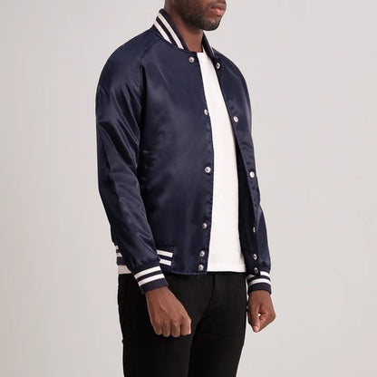 A man wearing a navy blue satin varsity-style bomber jacket with white and navy striped ribbed collar, cuffs, and hem, layered over a white t-shirt and black pants, photographed against a neutral light gray background.