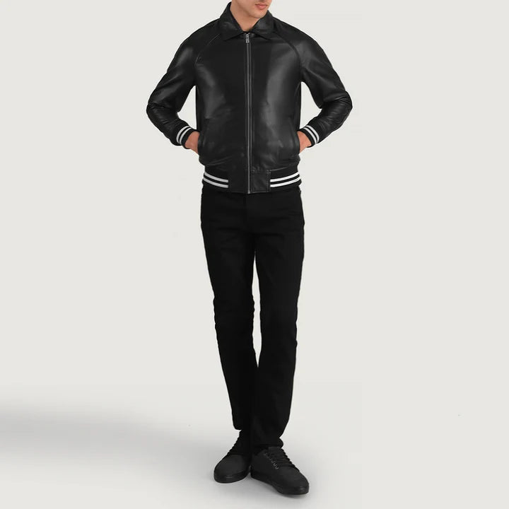 A man wearing a black leather bomber jacket with white stripes on the ribbed collar, cuffs, and hem, paired with black pants and black sneakers, set against a plain light gray studio background.