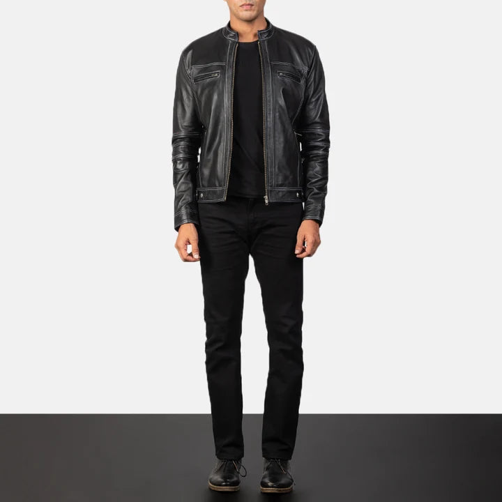A man wearing a black leather motorcycle jacket over a black t-shirt, black jeans, and black shoes, standing against a split white and dark gray studio background.