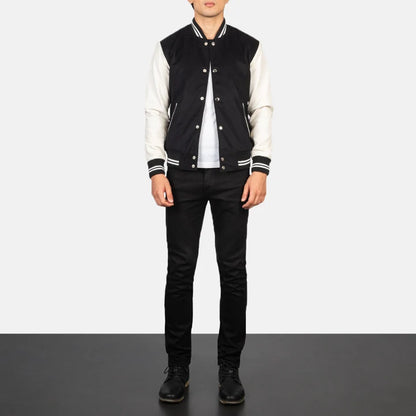 A model wearing a black and white varsity jacket, black jeans, and black shoes, standing against a split white and gray studio background.