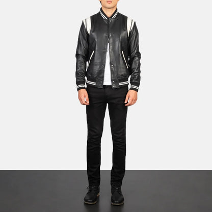 A man wearing a black leather varsity-style jacket with white striped ribbed collar, cuffs, and shoulders, paired with black jeans and black shoes, standing against a white studio background.